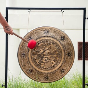 Peut inclure: Un grand gong orn&eacute; d'un motif d&eacute;taill&eacute; de dragon et de ph&eacute;nix. Le gong de couleur bronze est suspendu &agrave; un cadre en m&eacute;tal noir. Une personne tient une mailloche avec une t&ecirc;te de frappe rouge, pr&ecirc;te &agrave; jouer de l'instrument. Le gong est un instrument de musique.
