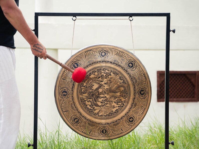 Peut inclure: Un grand gong orn&eacute; d'un motif d&eacute;taill&eacute; de dragon et de ph&eacute;nix. Le gong de couleur bronze est suspendu &agrave; un cadre en m&eacute;tal noir. Une personne tient une mailloche avec une t&ecirc;te de frappe rouge, pr&ecirc;te &agrave; jouer de l'instrument. Le gong est un instrument de musique.