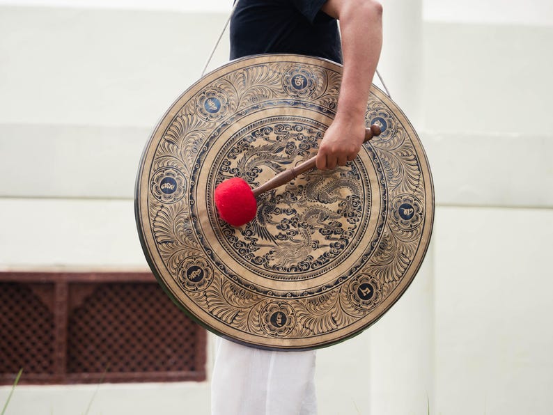 May include: A large, round gong with intricate engraved designs. The gong is primarily gold with dark brown detailing and a central dragon motif. A red mallet rests against the gong's surface. The gong is held by a person, suggesting its use in sound healing or musical performances.
