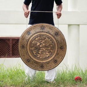 Peut inclure: Un grand gong rond de couleur bronze, orn&eacute; de gravures complexes de dragons et de motifs floraux. Le gong est suspendu par une corde et tenu par une personne v&ecirc;tue d'un t-shirt noir et d'un pantalon blanc. En arri&egrave;re-plan, de l'herbe verte et un mur blanc.