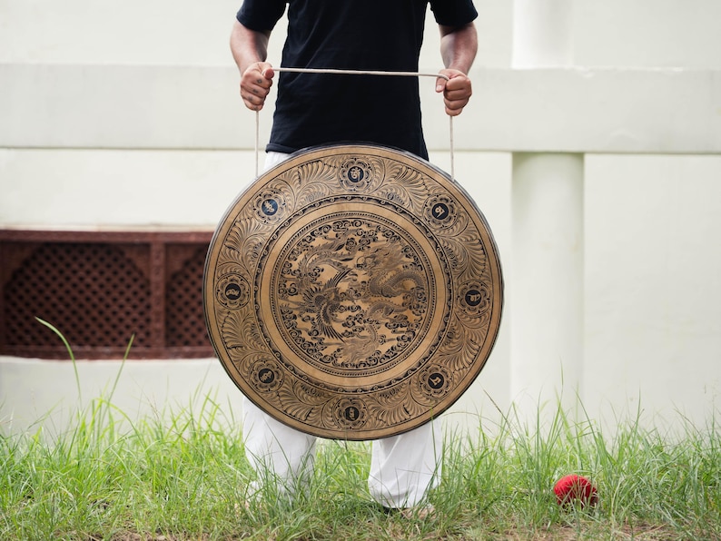 Peut inclure: Un grand gong rond de couleur bronze, orn&eacute; de gravures complexes de dragons et de motifs floraux. Le gong est suspendu par une corde et tenu par une personne v&ecirc;tue d'un t-shirt noir et d'un pantalon blanc. En arri&egrave;re-plan, de l'herbe verte et un mur blanc.