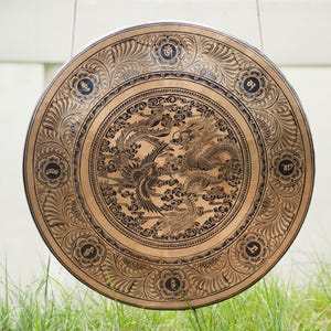 Peut inclure: Un grand gong circulaire avec un design orn&eacute;. Le gong est de couleur bronze avec des gravures noires complexes de dragons, de ph&eacute;nix et de motifs floraux. Le gong est suspendu par un cadre en m&eacute;tal noir, avec de l'herbe verte au premier plan et un fond blanc.