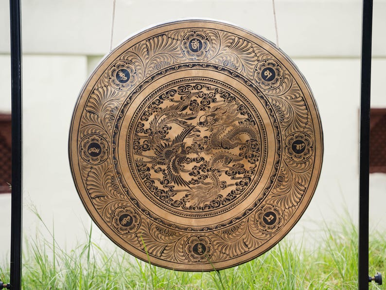 Peut inclure: Un grand gong circulaire avec un design orn&eacute;. Le gong est de couleur bronze avec des gravures noires complexes de dragons, de ph&eacute;nix et de motifs floraux. Le gong est suspendu par un cadre en m&eacute;tal noir, avec de l'herbe verte au premier plan et un fond blanc.