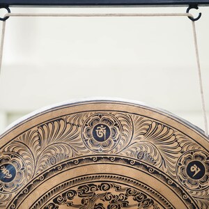 Peut inclure: Gros plan sur un gong d&eacute;coratif avec des motifs floraux noirs complexes sur une surface dor&eacute;e. Le gong est suspendu par des cordons blancs &agrave; une barre m&eacute;tallique noire. Le design comprend des symboles circulaires.