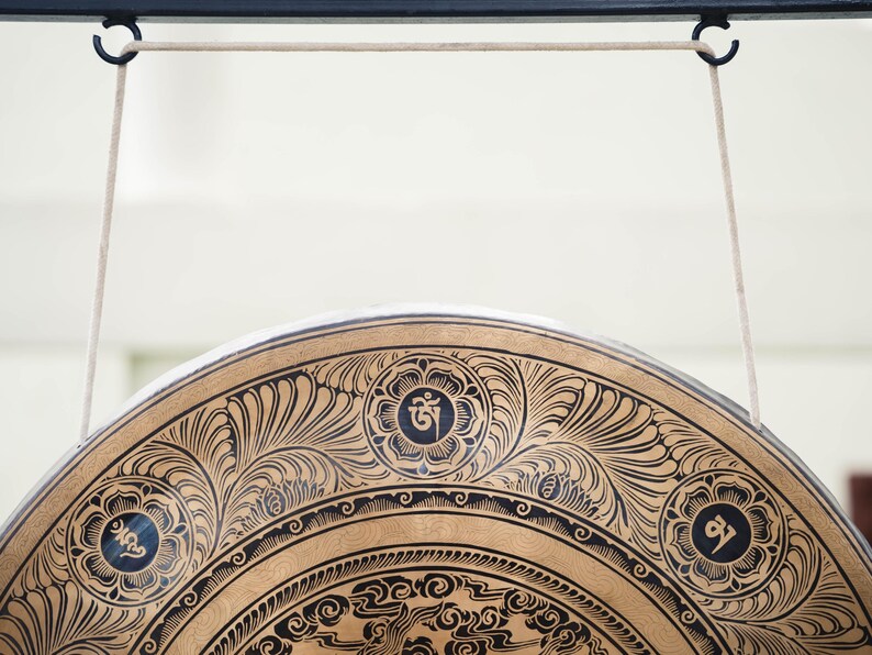 Peut inclure: Gros plan sur un gong d&eacute;coratif avec des motifs floraux noirs complexes sur une surface dor&eacute;e. Le gong est suspendu par des cordons blancs &agrave; une barre m&eacute;tallique noire. Le design comprend des symboles circulaires.