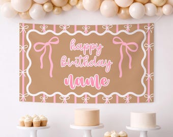 May include: A brown banner with pink text that reads "happy birthday name" is displayed. The banner has a white and pink border with pink bows. The background includes a balloon arch in shades of white, beige, and gold, and a table with cakes and cupcakes.