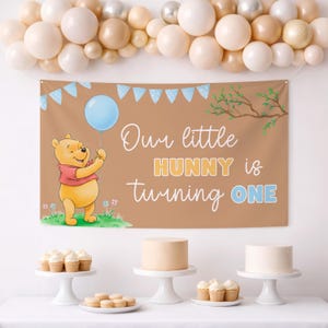 Può includere: Un banner di compleanno marrone con la scritta "Our little HUNNY is turning ONE" e un'illustrazione di Winnie the Pooh con un palloncino blu. La scena comprende un arco di palloncini e vari dolci, perfetto per un primo compleanno.