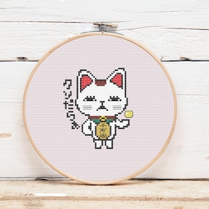 May include: Cross-stitch embroidery of a white cat with red ears and a gold coin, in a wooden hoop. The cat has a green and gold collar and holds a coin. Japanese text is on the left side of the cat.