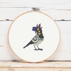 May include: A cross-stitch embroidery of a stylized bird in a wooden hoop. The bird features a black body, white accents, and a blue and yellow headpiece. The background is white, and the hoop is made of light-colored wood.