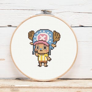 May include: A cross-stitch embroidery of a cartoon character, framed in a wooden hoop. The character has a blue hat with a pink cross, brown antlers, and a yellow and brown striped shirt. The background is white.