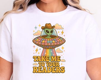 May include: White t-shirt featuring a retro-style graphic of an alien in a cowboy hat piloting a UFO. The UFO emits a rainbow beam, and the text "TAKE ME TO YOUR READERS" is below the UFO. The design includes clouds, stars, and lightning bolts.