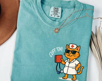May include: Teal Comfort Colors t-shirt featuring a cartoon orange cat dressed as a nurse, wearing sunglasses and holding a red box. The text "CUFF 'EM!" is above the cat. A gold necklace is draped across the shirt.