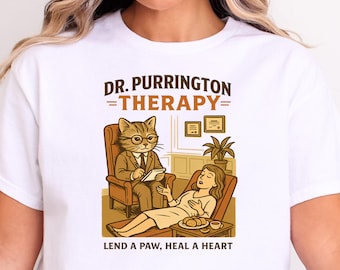 May include: White t-shirt featuring a cartoon illustration of a cat therapist in a suit and glasses, with a patient on a couch. The text reads "DR. PURRINGTON =THERAPY= LEND A PAW, HEAL A HEART."