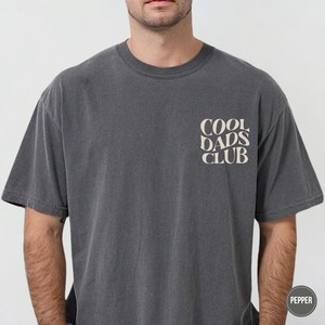 Man wearing a faded Pepper gray Comfort Colors t-shirt with a small cream Cool Dads Club graphic printed on the left chest.