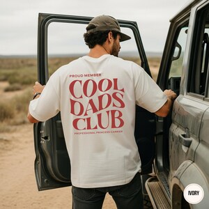 Back view of a man standing beside an off-road vehicle wearing an ivory t-shirt with a large red Cool Dads Club graphic and Professional Princess Carrier text.