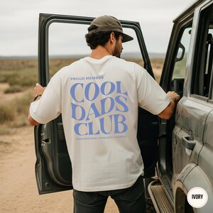 Back view of an ivory t-shirt with a large light blue Cool Dads Club design and Professional Princess Carrier text.