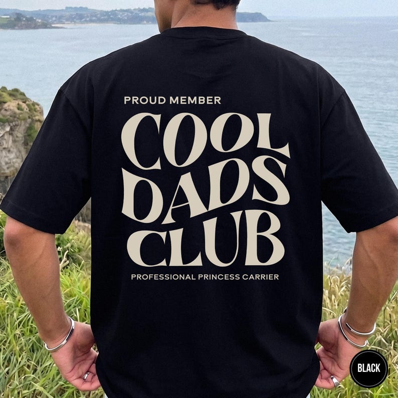 Man standing outdoors wearing a black t-shirt with a large cream Cool Dads Club graphic printed across the back.
