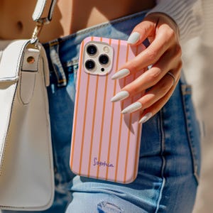 May include: A pink phone case with vertical orange stripes. The case has a camera cutout and the name "Sophia" in cursive. The phone case is held in a person's hand, with long, white fingernails.