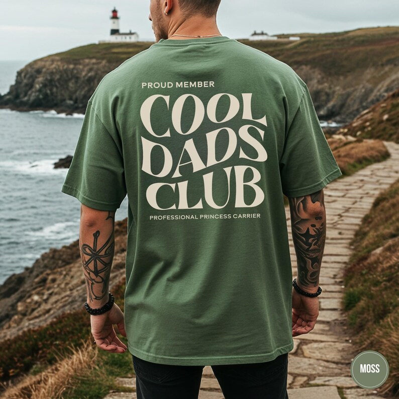 Man standing on a coastal path wearing a Moss green t-shirt with a large cream Cool Dads Club graphic on the back.