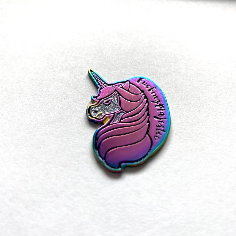 May include: A rainbow iridescent enamel pin featuring a unicorn head with a glitter horn and the text "Fucking Majestic".
