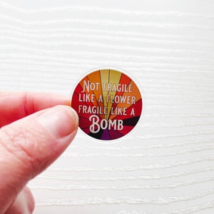 May include: A round sticker with a colorful, sunburst design. The sticker says "Not Fragile Like A Flower Fragile Like A Bomb".