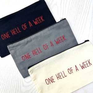 May include: Three zippered pouches with the text "ONE HELL OF A WEEK" embroidered in red on each pouch. The pouches are made of different fabrics: black, gray, and white.