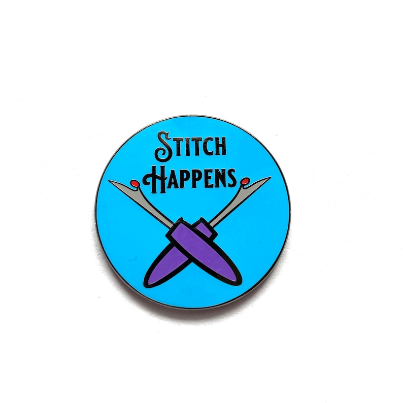 Pu&ograve; includere: Una spilla rotonda blu chiaro con la scritta "Stitch Happens" in nero. Due taglia-cuci viola incrociati sotto. La spilla ha un bordo argentato e dettagli rossi.