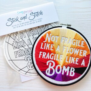 以下が含まれることがあります： 虹色の文字「Not Fragile Like A Flower Fragile Like A Bomb」が刺繍された完成した刺繍枠のクローズアップ。枠は白の表面にあり、黒と白のパターンが描かれた空白の刺繍枠の隣にあります。