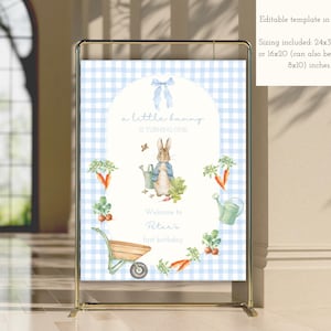 May include: A blue and white gingham patterned sign with the text "a little bunny is turning one." The sign features a watercolor illustration of a bunny, carrots, and a watering can. The sign is displayed on a gold metal stand.