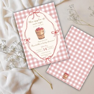 May include: Baby shower invitations with a pink and white gingham pattern. The main invitation features an oval frame with a watercolor illustration of a wooden bucket filled with apples and the text "the apple of our eye is on the way".