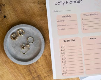 Puede incluir: Un planificador diario con un diseño melocotón claro y blanco, con secciones para programar, seguimiento de agua, listas de tareas y notas. Una bandeja de hormigón gris contiene pendientes y anillos dorados. Los artículos descansan sobre una superficie de madera.