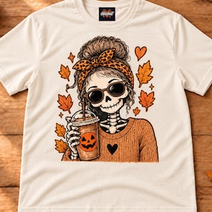 Op de afbeelding: Een crèmekleurig T-shirt met korte mouwen en een afbeelding van een skelet met een haarband met luipaardprint en een zonnebril, die een ijskoffie met een pompoengezicht vasthoudt. Het ontwerp bevat oranje herfstbladeren en een hartje.