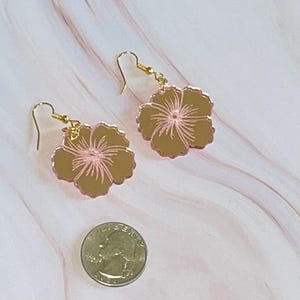 Puede incluir: Un par de pendientes dorados con forma de flores de hibisco. Los pétalos de la flor son de color marrón claro con detalles en rosa. Los pendientes cuelgan de ganchos dorados. Una moneda está en primer plano.