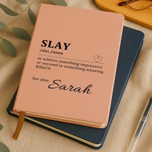 Może przedstawiać: Różowy notatnik z napisem "SLAY" i jego definicją na okładce, wraz z imieniem "Sarah". Notatnik leży na ciemnoniebieskim notatniku, z długopisem i okularami w tle.