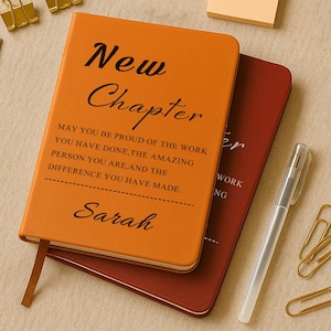 May include: An orange journal with the words "New Chapter" and a personalized message printed in black. A maroon journal is partially visible beneath it. A pen, paperclips, and a sticky note are also present.