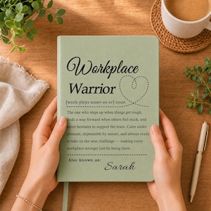 Könnte beinhalten: Ein salbeigrünes Notizbuch mit dem schwarz aufgedruckten Text Workplace Warrior, einer Definition des Begriffs, einer gestrichelten Herz-Kontur und dem Namen Sarah unten. Der Einband hat eine glatte, matte Oberfläche und wird von zwei Händen gehalten.