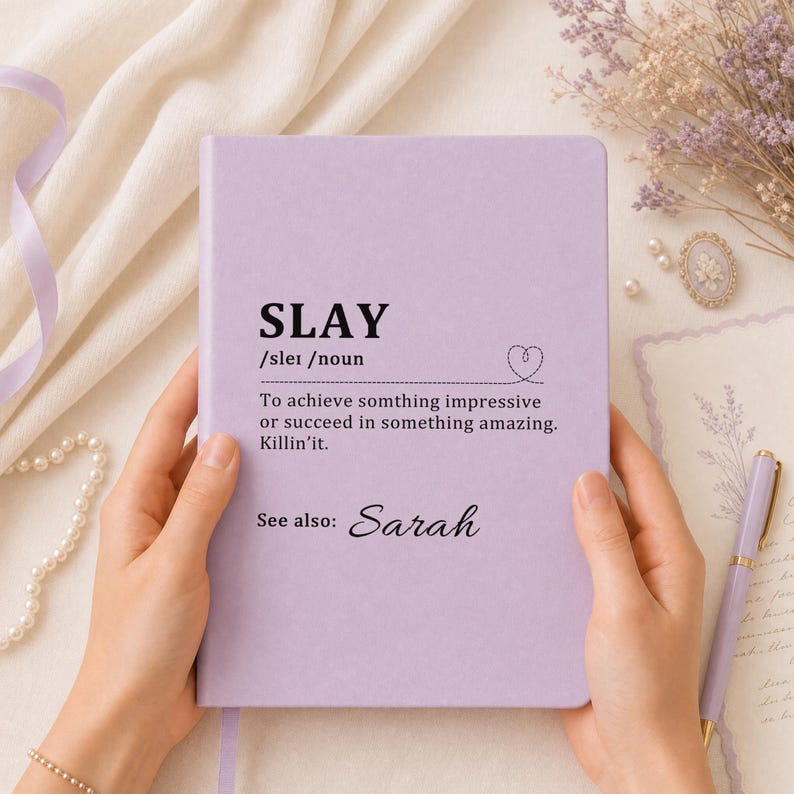 May include: A light purple notebook with black text that reads SLAY /slei /noun To achieve somthing impressive or succeed in something amazing. Killin'it. See also: Sarah. The cover features a small dashed heart icon next to the definition.
