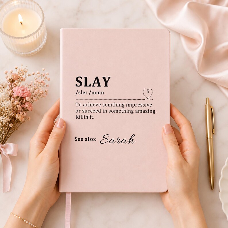 May include: A light pink notebook featuring the black printed text SLAY /slei /noun, a definition of the word, a small heart icon, and the text See also: Sarah. The cover has a smooth, matte finish and is held upright in a vertical orientation.
