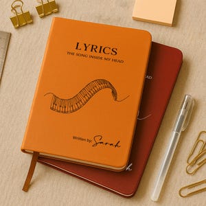 May include: An orange notebook with the word "LYRICS" and the phrase "THE SONG INSIDE MY HEAD" printed on the cover. A piano keyboard design is below the text. The notebook is stacked on top of a burgundy notebook. A pen and paper clips are also in the image.