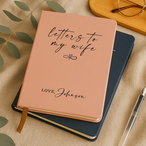 May include: A pink journal with the text "Letters to my wife" and "Love, Johnson" in black script. The journal has a gold ribbon bookmark and rests on top of a dark blue journal.