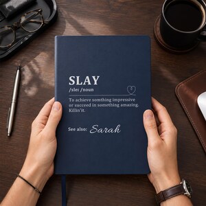 May include: A dark blue notebook with white text that reads SLAY /sleɪ /noun To achieve somthing impressive or succeed in something amazing. Killin'it. See also: Sarah. The cover features a small heart icon and is held by two hands.