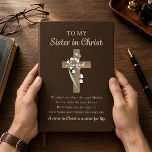 May include: A dark brown journal featuring the gold text TO MY Sister in Christ, a wooden cross graphic with lily of the valley flowers, the name Sarah, and the poem: We might not share the same Mother but we share the same Father He brought you into my life for a reason and I thank Him every day A sister in Christ is a sister for life.