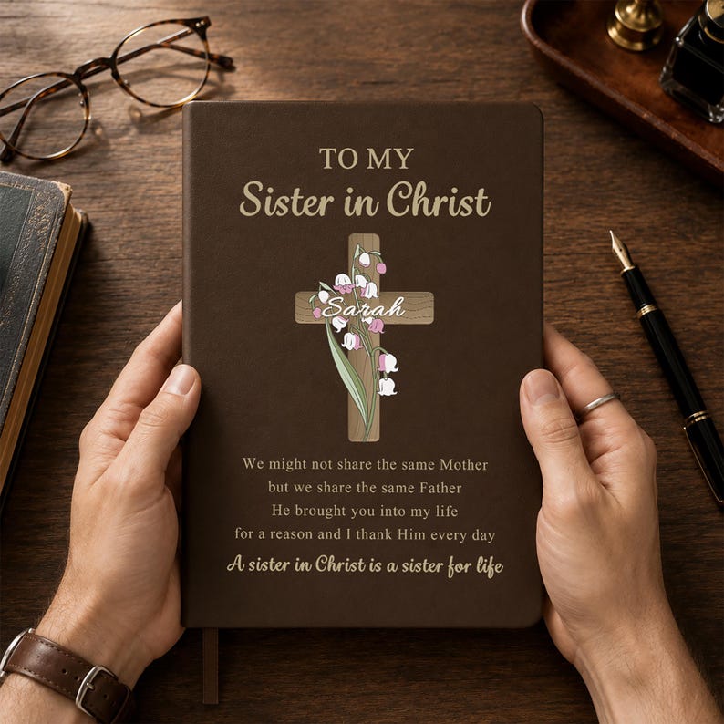 May include: A dark brown journal featuring the gold text TO MY Sister in Christ, a wooden cross graphic with lily of the valley flowers, the name Sarah, and the poem: We might not share the same Mother but we share the same Father He brought you into my life for a reason and I thank Him every day A sister in Christ is a sister for life.
