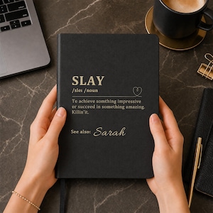 May include: A black hardcover notebook featuring gold-colored text that defines the word SLAY as achieving something impressive or succeeding in something amazing, with the name Sarah printed below as a reference, all in a clean, professional layout.