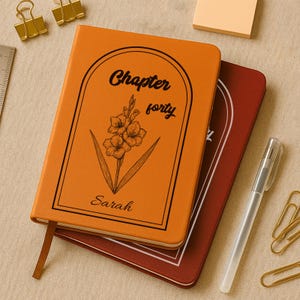 Può includere: Un diario arancione con le parole "Chapter forty" e il nome "Sarah" stampati sulla copertina, insieme a un'illustrazione floreale. Sotto è visibile un secondo diario. Sono presenti anche una penna, dei post-it e delle graffette.