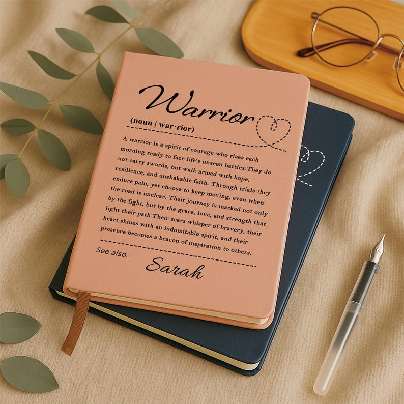 Peut inclure: Un carnet couleur p&ecirc;che avec la d&eacute;finition imprim&eacute;e en noir du mot Warrior et le pr&eacute;nom Sarah. Le carnet pr&eacute;sente une petite ic&ocirc;ne en forme de c&oelig;ur et un marque-page en ruban marron. Il est pos&eacute; sur un carnet bleu fonc&eacute;, accompagn&eacute; d'un stylo-plume.
