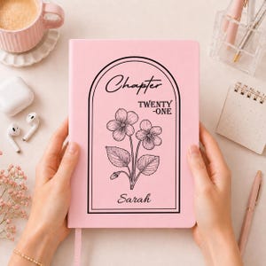 May include: A light pink notebook featuring a black arched frame design on the cover. Inside the arch, the text reads Chapter TWENTY-ONE above a black line drawing of a flower, with the name Sarah printed in black script at the bottom.