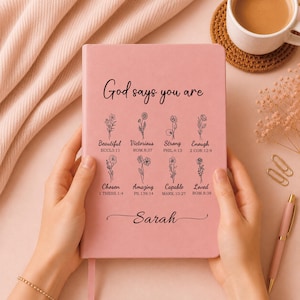 Può includere: Un taccuino rosa con il testo God says you are, seguito da otto illustrazioni floreali con parole e versetti biblici corrispondenti, e il nome Sarah stampato in basso in un carattere corsivo. La copertina ha una finitura liscia e opaca.