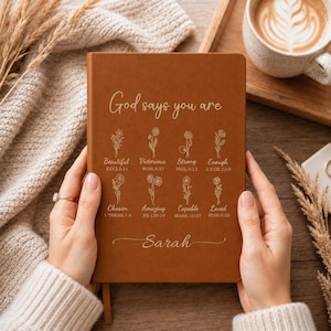 Peut inclure: Un carnet marron avec du texte en feuille d'or indiquant God says you are, suivi de huit illustrations florales avec des étiquettes comme Beautiful, Victorious, Strong, Enough, Chosen, Amazing, Capable, Loved, et le nom Sarah en bas.