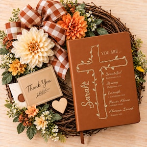 May include: A brown leather-bound journal featuring a vine-wrapped cross and the text "YOU ARE... Beautiful, Strong, Enough, Never Alone, Always Loved" with scripture references, resting on a wreath with orange flowers and a plaid ribbon.